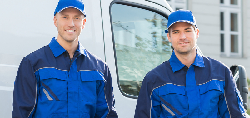 Two men in blue work clothes standing next to a white van parked on a street.